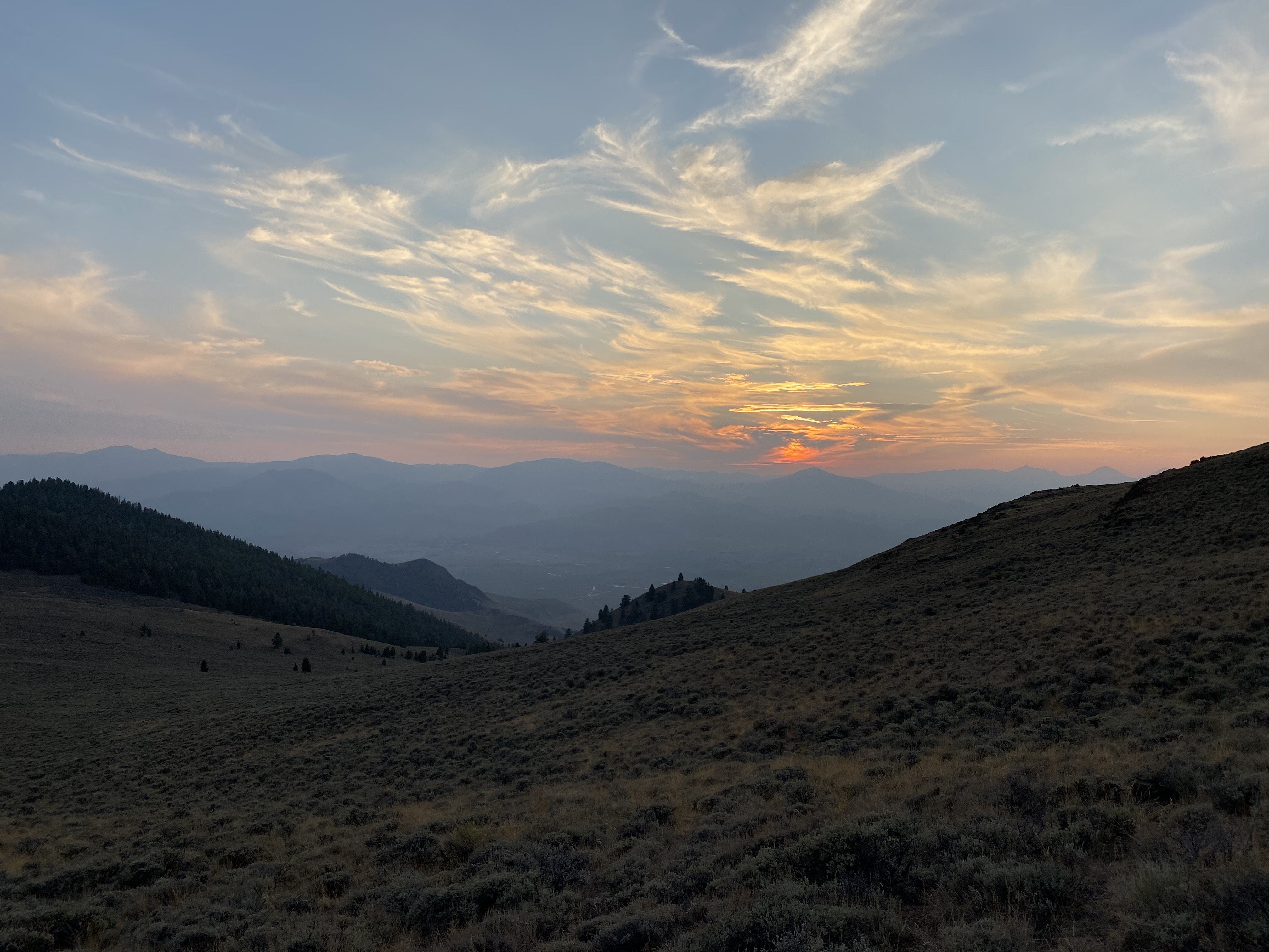 A sagebrush hillside at sunset looking out over layered mountain ridges and a valley, the sun breaking through streaked clouds on the horizon